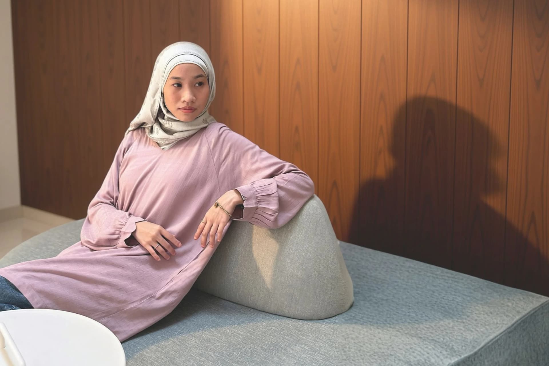 A woman in the lavender Anthea long dress and a cream headscarf, leaning on a grey sofa